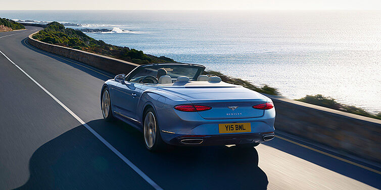 Bentley Rotterdam New Bentley Continental GTC Azure convertible rear view in Light Windsor blue paint with 22 inch ten spoke wheels driving dynamically on an ocean road in the sunshine